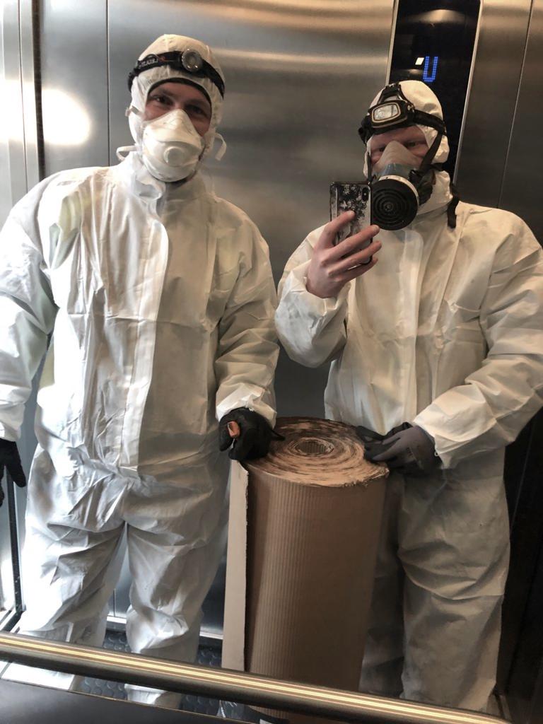Kaksi suojahaalariin, hengityssuojaimiin ja suojakäsineisiin pukeutunutta remonttimiestä ottaa hyväntuulista selfietä hississä. Miehet nojaavat suojapaperirullaan.