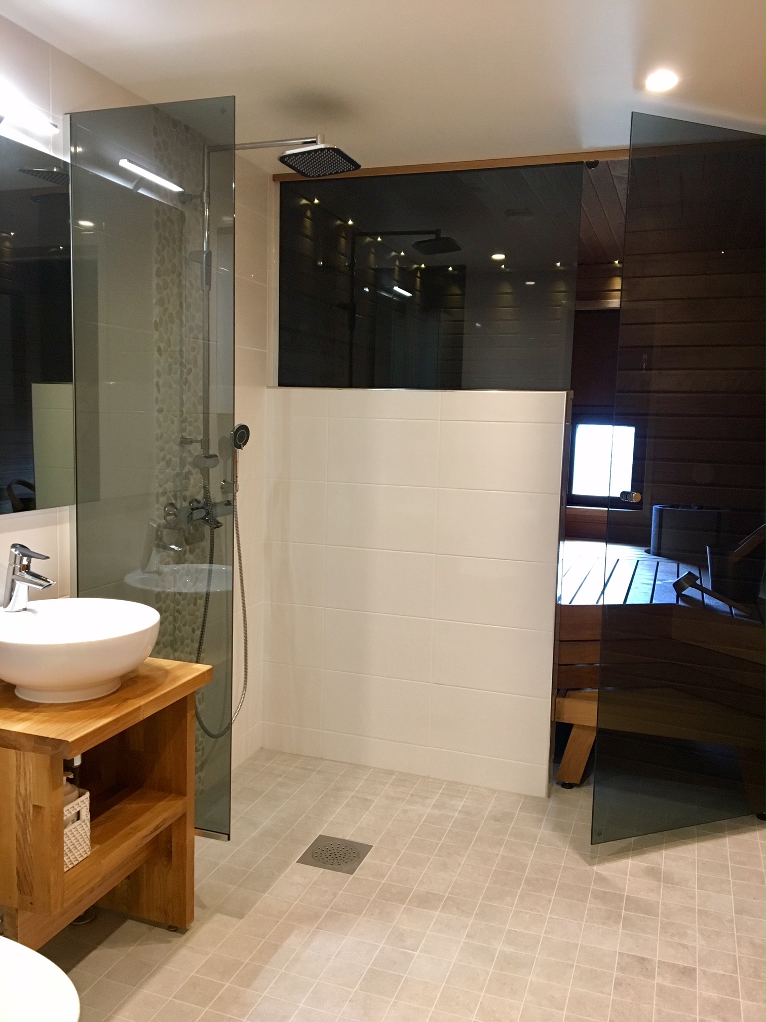 Raikas, vastaremontoitu kylpyhuone. Etualalla pyöreä käsienpesuallas ja käsintehty allastaso. Taustalla suihkunurkkaus, jossa koristekivilaatoitettu seinä ja isot vaakalaatat. Taaimmaisena sauna, josta tummennettu ikkuna suihkutiloihin.
